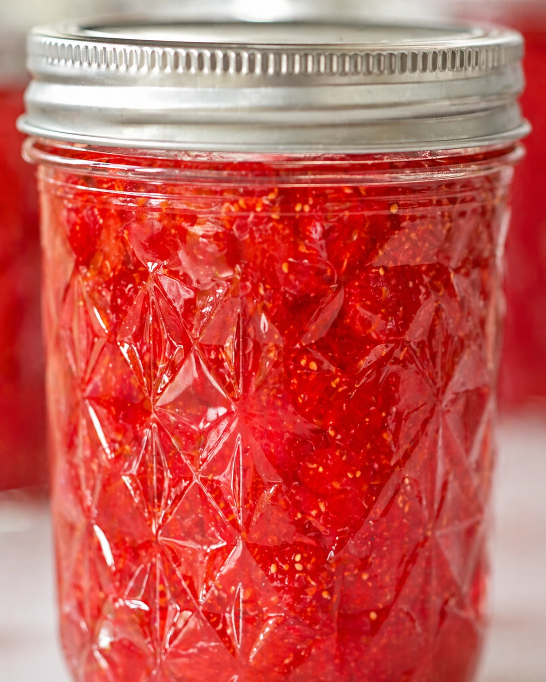 Close-up of mason jar showing chunky ruby-red strawberry jam through embossed glass.